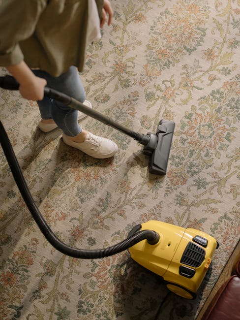 Close-up of a person kneeling on a patterned carpet, using a yellow and black vacuum cleaner for surface cleaning. The individual is handling a white cloth and placing it inside the vacuum's dust comp